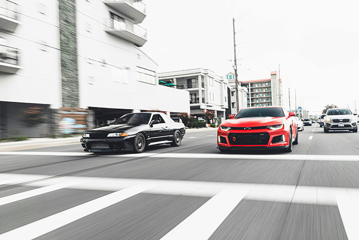 Two powerful cars, a black Nissan Skyline and red Chevy Camaro, driving fast on a road, a scene suggesting road rage.