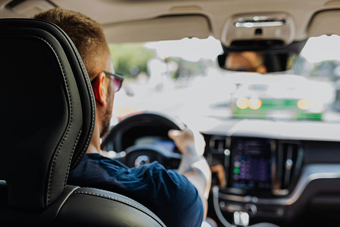 A driver with sunglasses and tattoos, seen from behind, grips the steering wheel, indicating potential road rage.