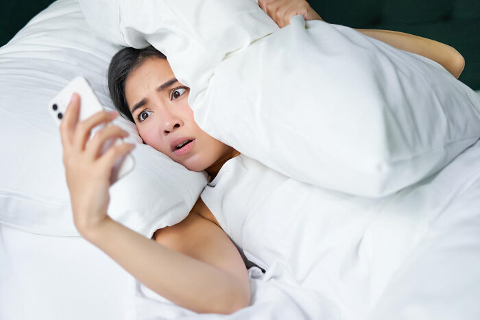 Worried woman hiding under covers looking at phone in bed