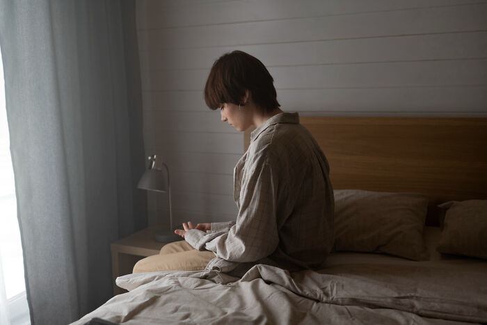 Sad person sitting on bed in dimly lit room looking down