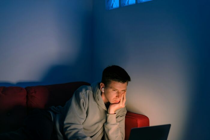 A young man in a grey hoodie, wearing earbuds, looking tired while using a laptop in a dimly lit room. He is one of the men suffering.