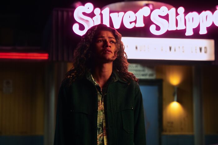 Zendaya em euforia, olhando para cima com uma expressão preocupada, na frente de um letreiro de néon. Fãs divididos pelo momento de Sydney Sweeney.