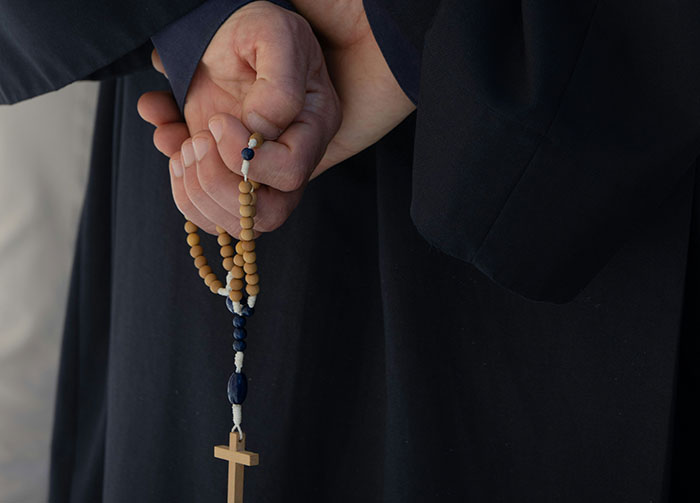 Hands clasped behind back, holding a rosary with wooden beads and a crucifix, symbolizing controversial beliefs.