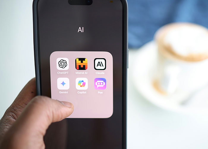 A hand points to a smartphone screen displaying an AI app folder, with icons for ChatGPT, Mistral AI, Claude, Gemini, Copilot, and Poe. Controversial beliefs about AI.