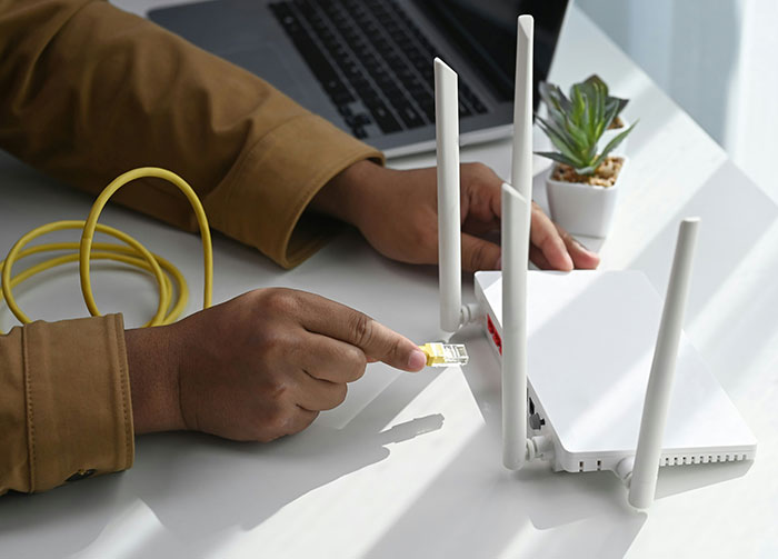 Person in brown shirt connecting a yellow ethernet cable to a white router, with a laptop and plant in background. Might start arguments about tech setup.