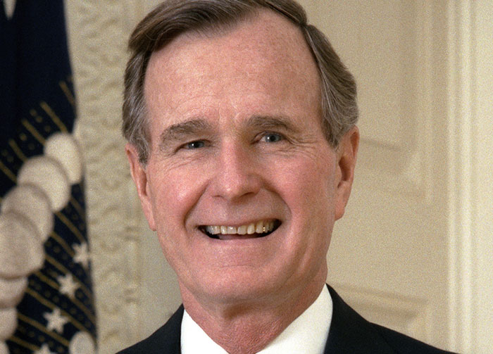 George H.W. Bush smiling, controversial beliefs, standing with a flag in the background, possibly starting arguments.