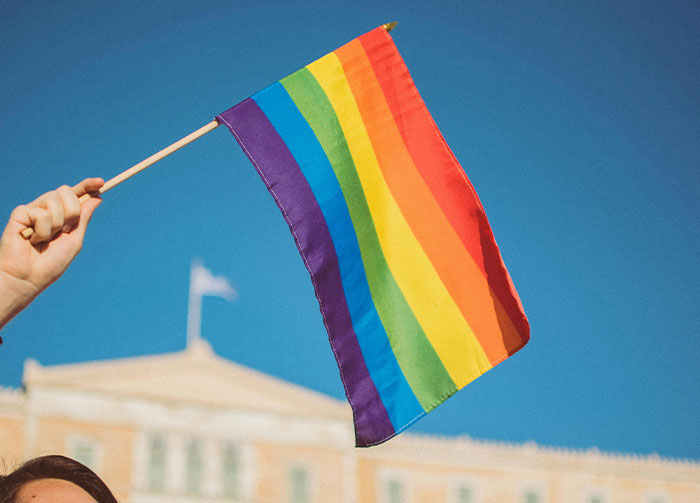 Hand holding a small rainbow flag against a clear blue sky, a building visible in the background. Controversial Beliefs.