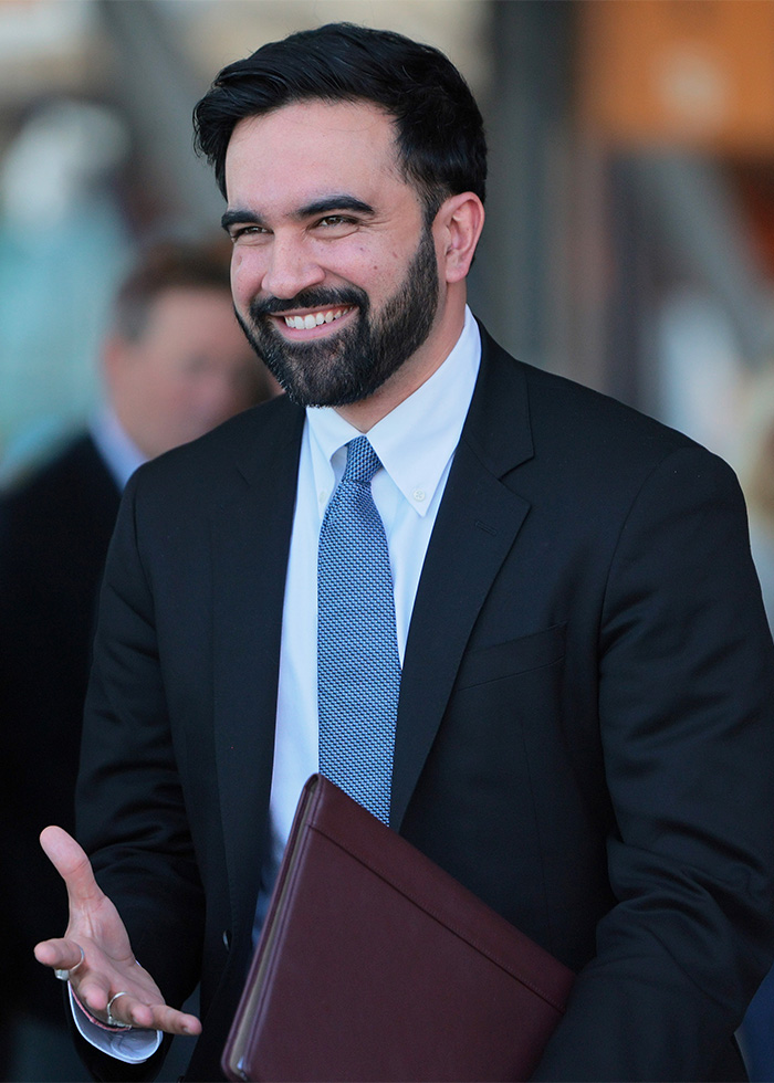 A smiling, bearded man in a suit and tie, holding a folder. Met Gala 2026 and Jeff Bezos scandals.
