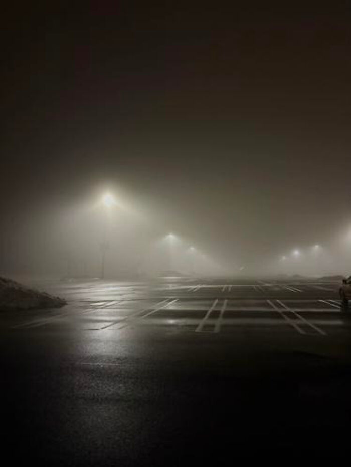 A vast, empty parking lot shrouded in dense fog with distant streetlights, a haunting liminal space.