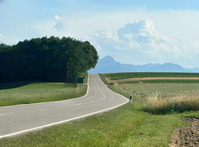 A winding road through green fields leading towards distant mountains under a cloudy sky, a natural liminal space.