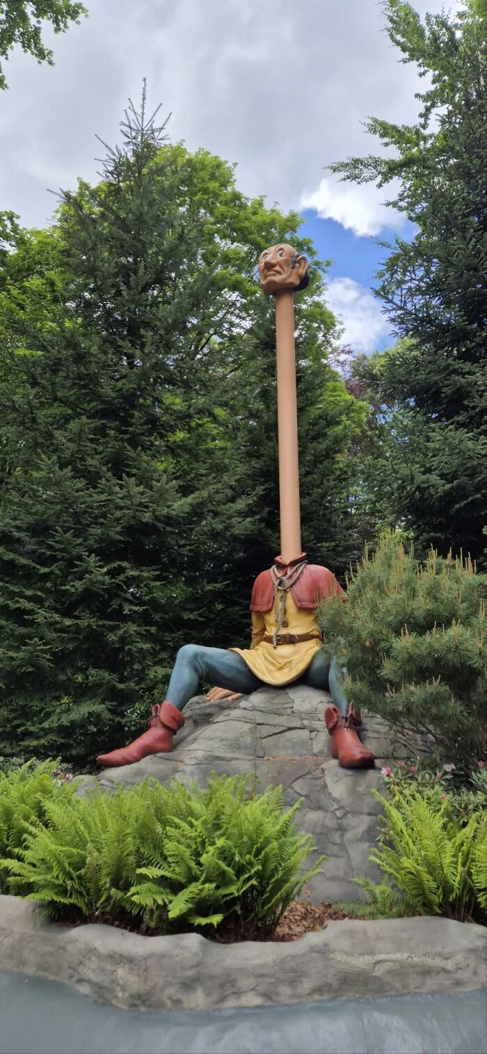 An image of a bizarre statue with a long neck and small head, sitting on a rock amidst trees.