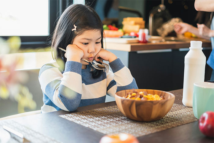 A young girl looking sad at breakfast, with a bowl of cereal, experiencing a food issue.