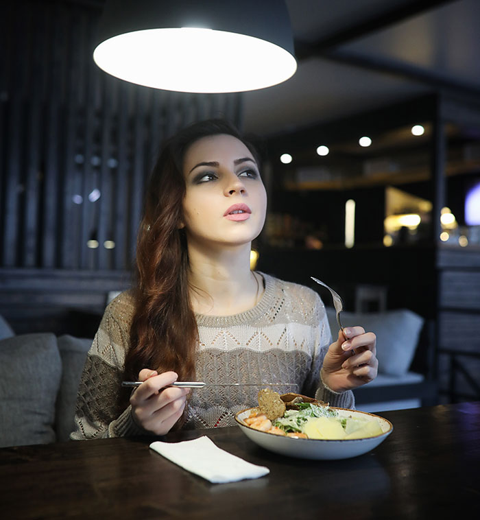 Woman looking upset while holding fork and knife eating meal under a lamp Woman looking upset while holding fork and knife eating meal under a lamp