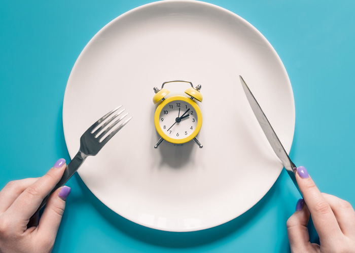 Fork and knife poised over plate with yellow alarm clock symbolizing fasting time Fork and knife poised over plate with yellow alarm clock symbolizing fasting time