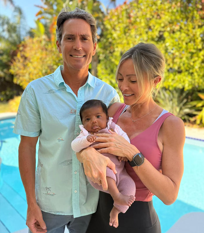 A couple smiling by a pool, holding the baby they gave birth to after an IVF mix-up, keeping her after a deal. A couple smiling by a pool, holding the baby they gave birth to after an IVF mix-up, keeping her after a deal.