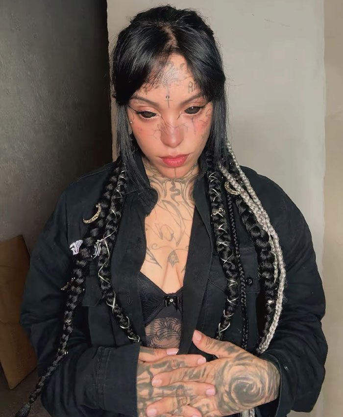 A woman with black tattooed eyes and numerous tattoos on her body, wearing a black open shirt and black bra, looking down thoughtfully.