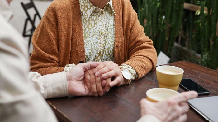 An elderly couple holding hands on a date, sitting at a wooden table with coffee cups and a smartphone.