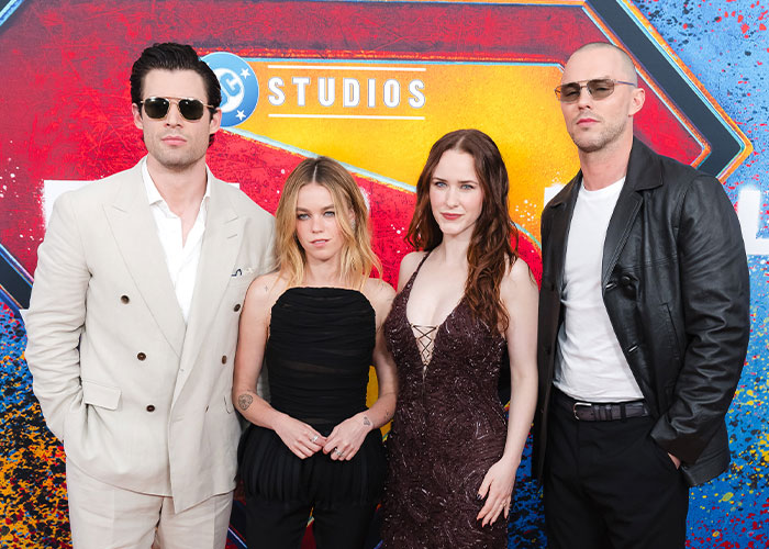 The cast of the Supergirl movie standing on a red carpet with a colorful background, posing for cameras.
