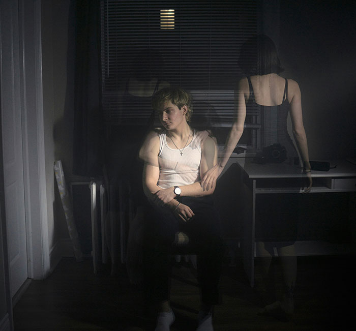 A man sitting in a dimly lit room, with a translucent female figure behind him, representing the husband's stalkerish tendencies. A man sitting in a dimly lit room, with a translucent female figure behind him, representing the husband's stalkerish tendencies.