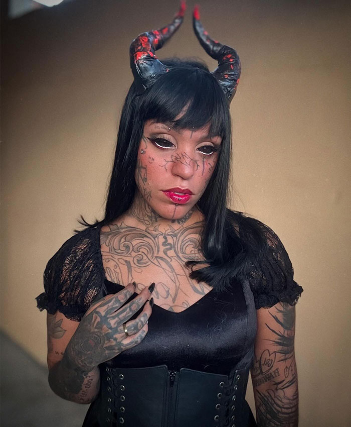 A woman with tattooed eyes and horns on her head, wearing a black top with lace sleeves and a corset, poses for the camera.