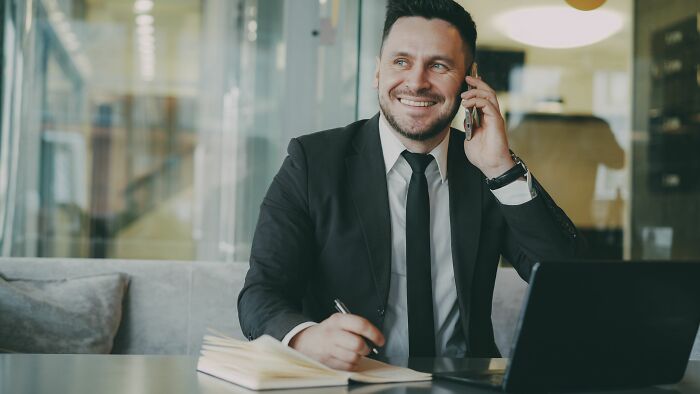 A smiling man in a suit talks on the phone while working on a laptop, showing acceptable actions on a date.