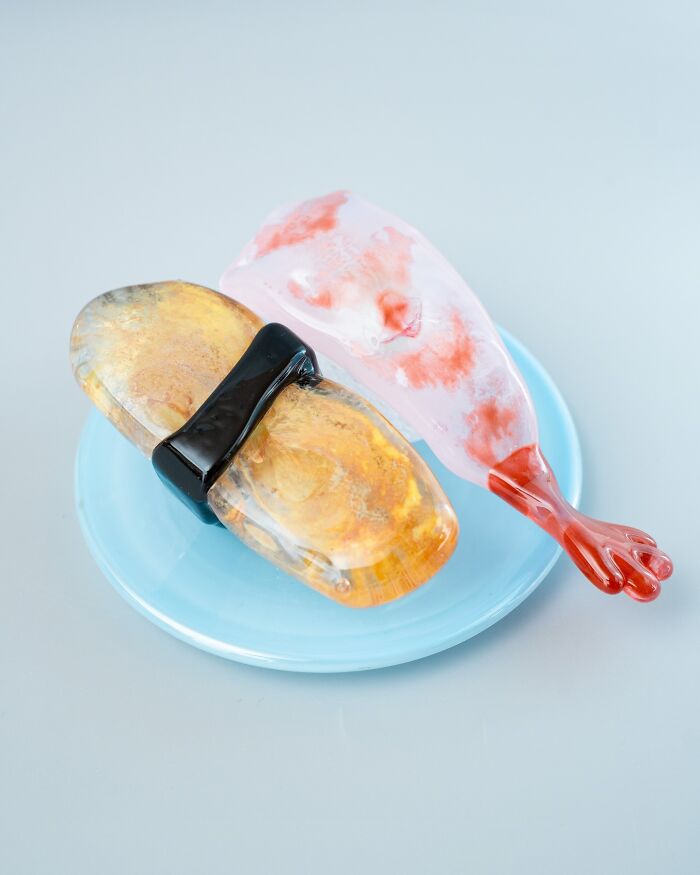 A close-up of hyper-realistic glass food sushi, an artist's creation that looks good enough to eat on a light blue plate.