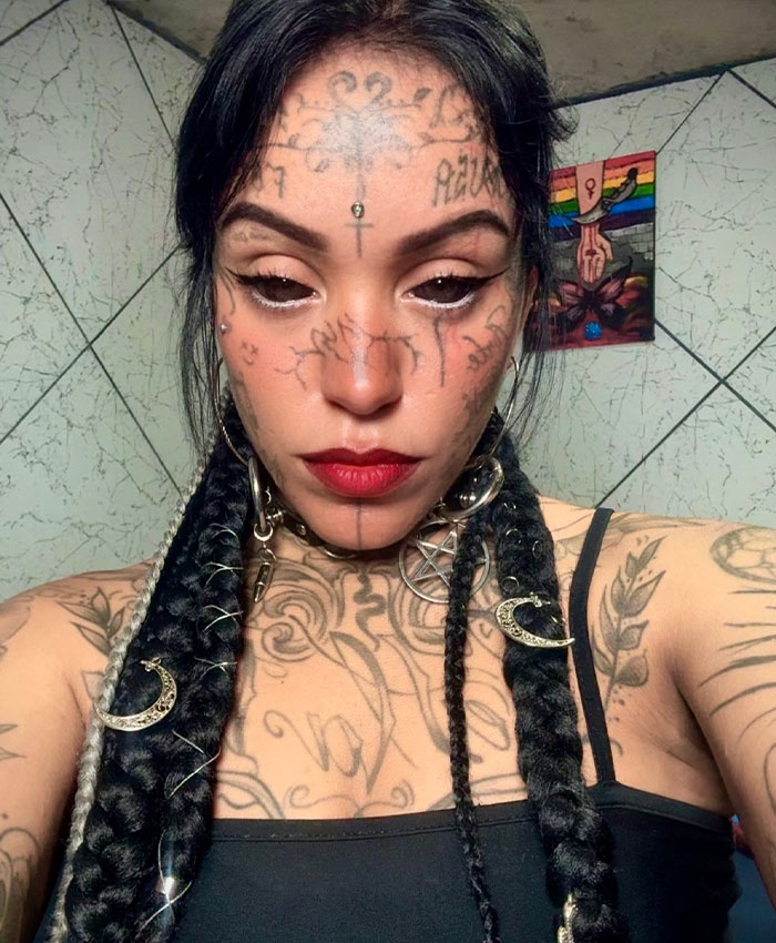 A woman with tattooed face, black tattooed eyes, and long braided hair with white accents, wearing a black tank top and metallic choker.