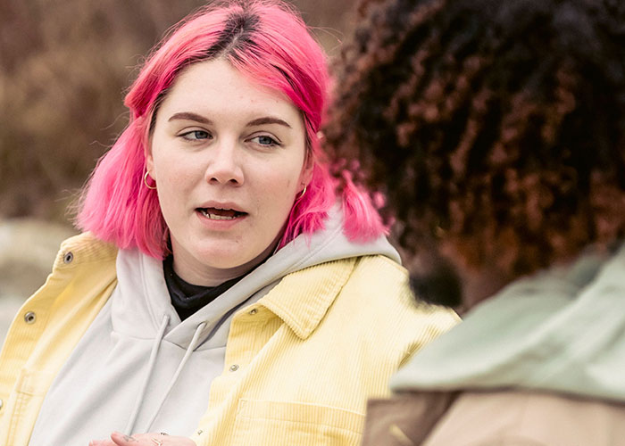 A woman with pink hair talking to a man, discussing how close friends broke trust.