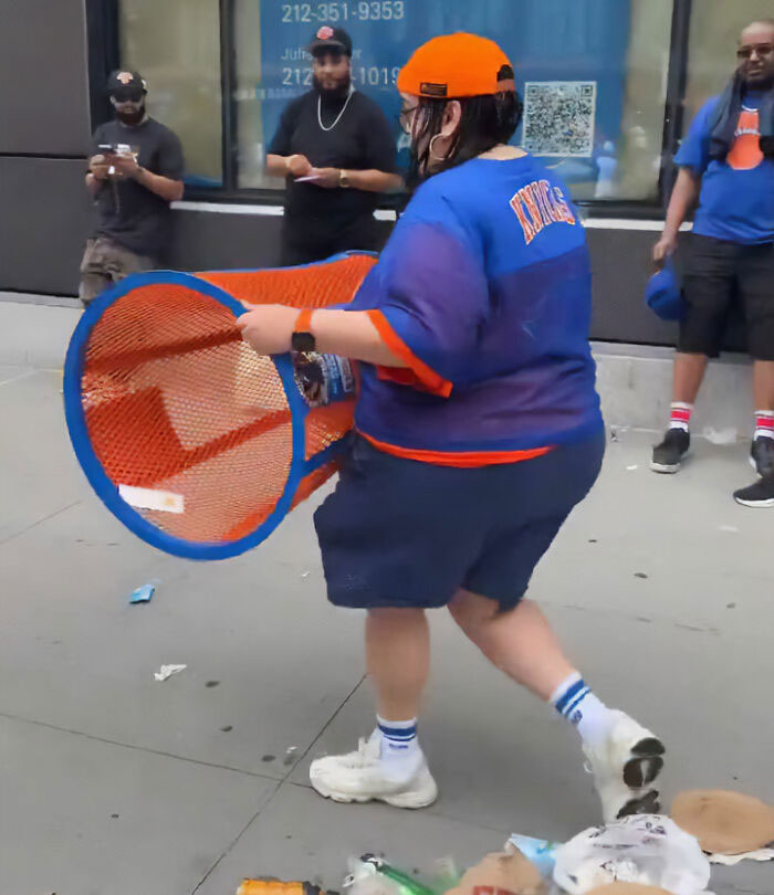A person in a Knicks jersey and orange hat carrying a stolen Knicks trashcan on a city street, part of a viral video.