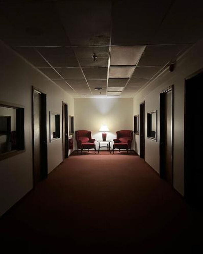 A dimly lit hotel corridor with two red armchairs and a lamp, creating a haunting liminal space.
