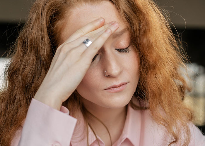 A young woman with red hair looking stressed, touching her forehead, realizing she can’t keep contact with her parents.