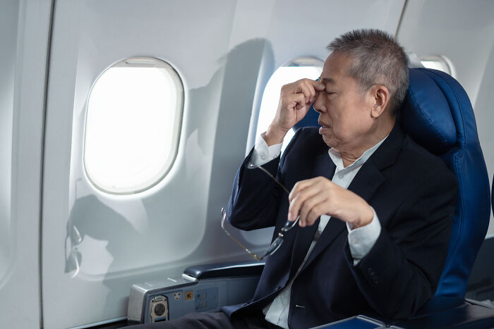 A man on a plane with his eyes closed and hand on his forehead, looking stressed. Craziest plane interactions.