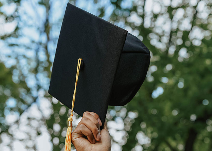 Hand holds up a graduation cap against a blurred green background, representing the post-high school life of popular kids.