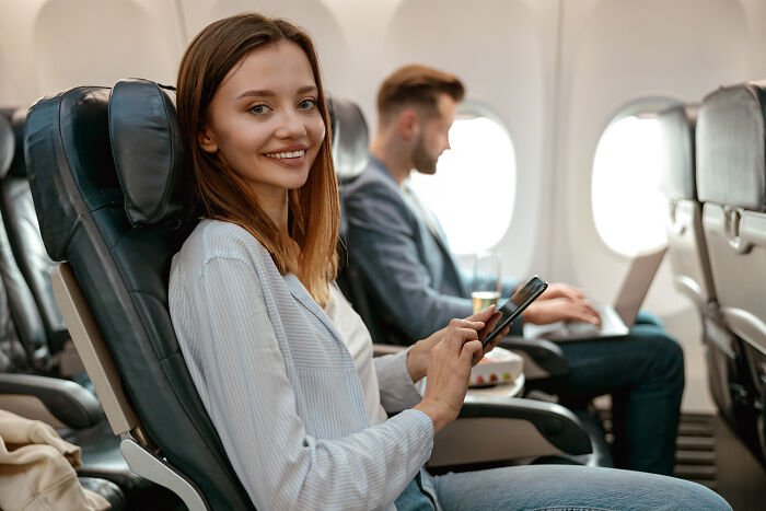 A woman smiles while using her phone on a plane, amidst various plane interactions.