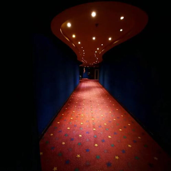 A long, dark hallway with a red, star-patterned carpet and bright overhead lights, a haunting liminal space.
