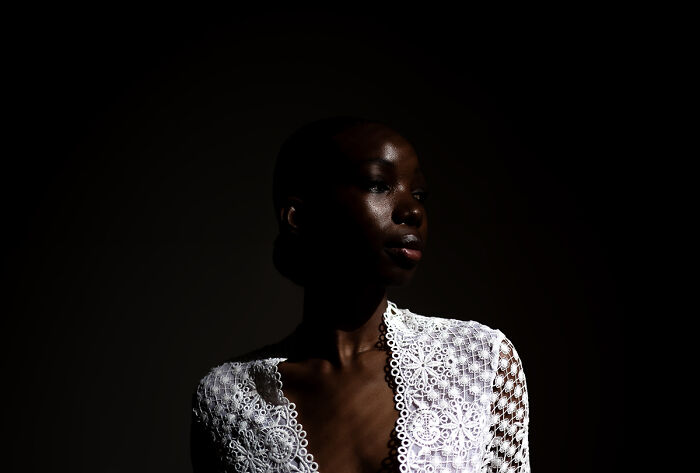 A woman with dark skin wears a white lace top, with light highlighting her face, a photo from the 2025 Chromatic Awards.