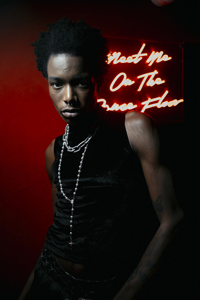 A person with dark skin and a nose ring wears a black top and silver necklaces, illuminated by neon, Chromatic Awards photo.