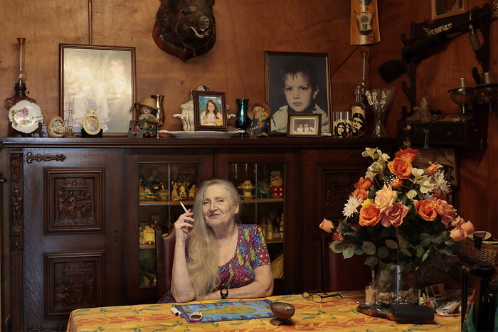 An older woman with long blonde hair sits at a table with flowers and photos, a compelling image from the Chromatic Awards.