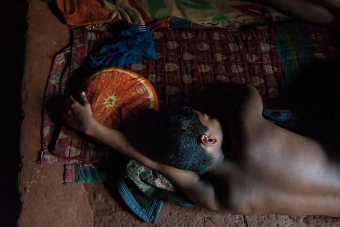 A person sleeps on a thin mattress with a round orange pillow, a poignant photograph from the Chromatic Awards.
