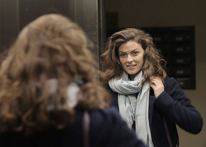 A young woman in a scarf and coat looking at her reflection in a mirror, possibly an elevator. She displays a serious problem-solving look.