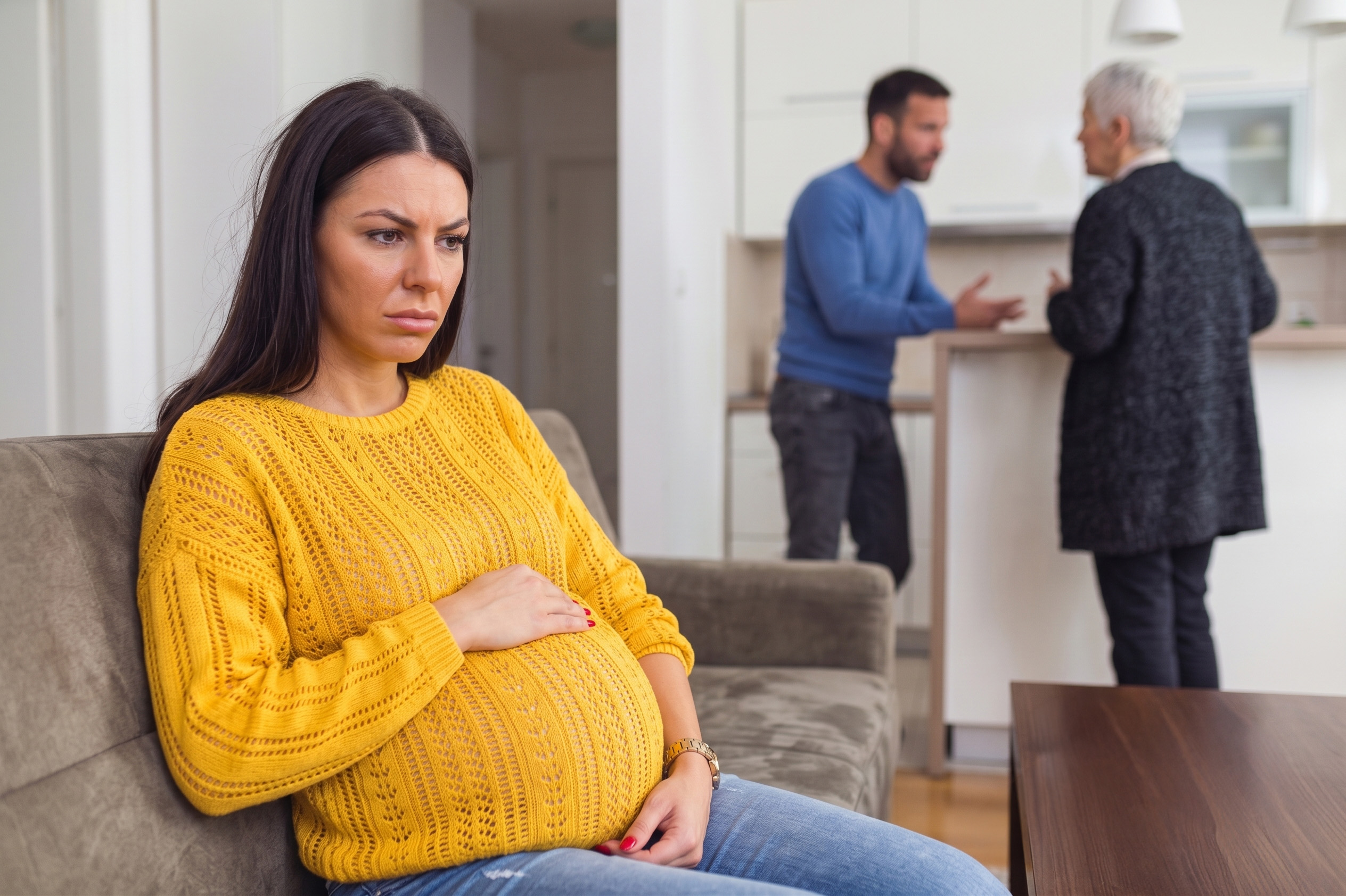 A pregnant wife, visibly upset, sits on a couch while her husband argues with his mom in the background, highlighting the husband wanting to honor his mom.