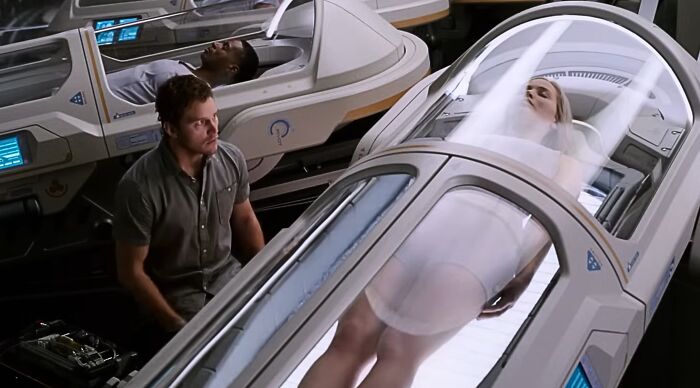 A man observes a woman in a hibernation pod, both characters from a popular fiction movie. The scene emphasizes the sci-fi setting.