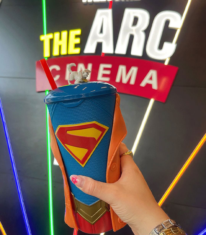 Supergirl fans hold a movie souvenir Grippr cup with a Supergirl design and a small dog figure at the cinema.