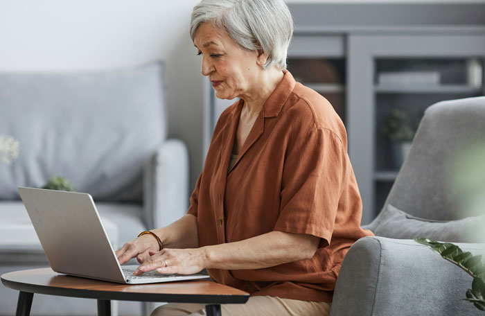 An elderly woman with gray hair working on a laptop in a living room, reflecting on heartwarming and heartbreaking things from late parents' phones.