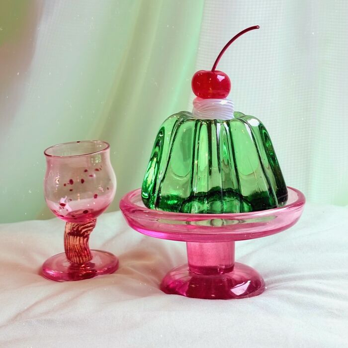 A green hyper-realistic glass food jelly with a cherry on top, next to a small pink glass.