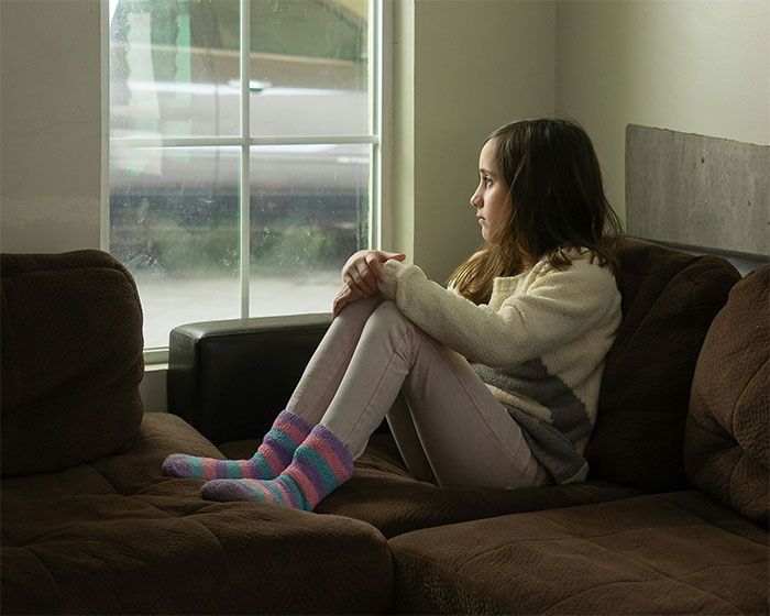 A young girl sitting on a couch, looking out a window, regretting a food issue situation.