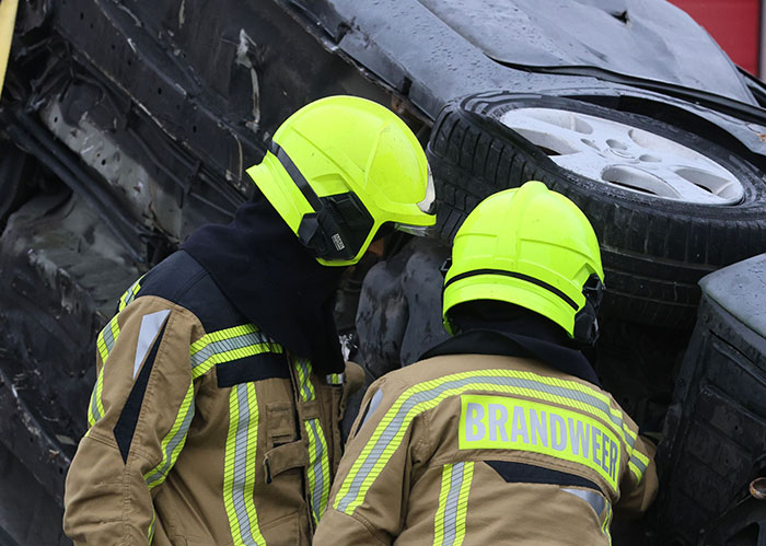 Two firefighters in neon helmets inspect a car accident scene, highlighting challenges after peaking in high school.