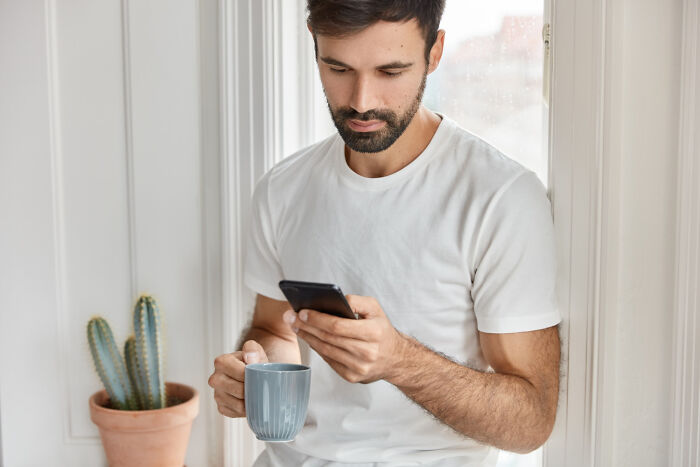A man looking at his phone, holding a cup, possibly reading about disturbing things people learned about their friends.