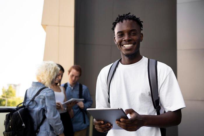 A smiling male student holding a tablet with friends in the background, depicting learning about friends.