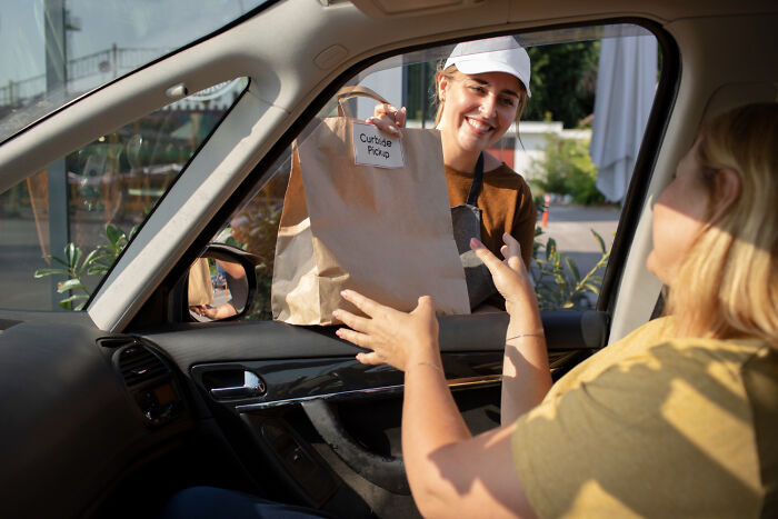 A smiling woman hands a curbside pickup bag to a customer in a car, representing disturbing things people learn about friends.